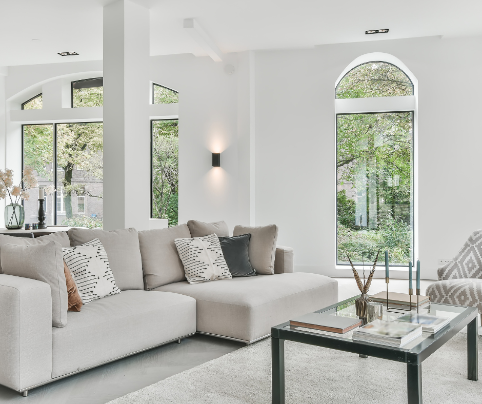 Bright, airy living room featuring a soft beige sectional sofa with neutral and patterned throw pillows, a glass coffee table with books and decor, and large arched windows letting in abundant natural light, surrounded by lush green outdoor views.