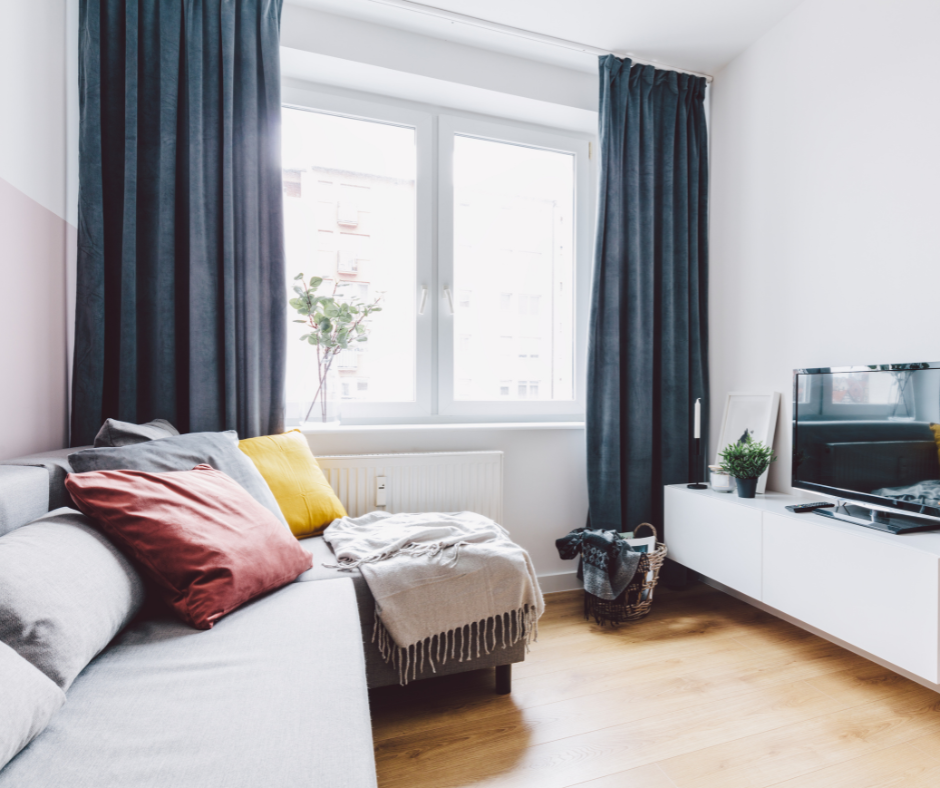 Small modern living room with high-hung dark curtains, gray sectional sofa, and bright natural light. Perfect example of how to make a small room look bigger with curtains and minimal decor.