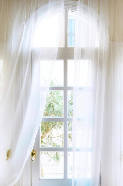 Bright doorway with arched window and sheer white curtains blowing gently in the breeze. Soft natural light and airy textures help make the room feel open and spacious.