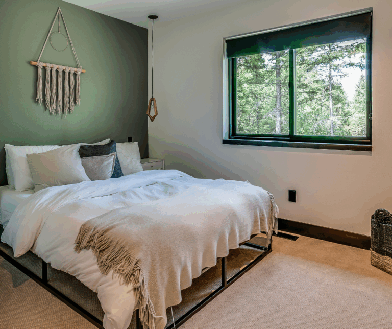 Cozy bedroom with a green accent wall behind the bed, featuring white bedding, neutral pillows, and a woven wall hanging. Large window with a view of pine trees outside adds natural light to the space.