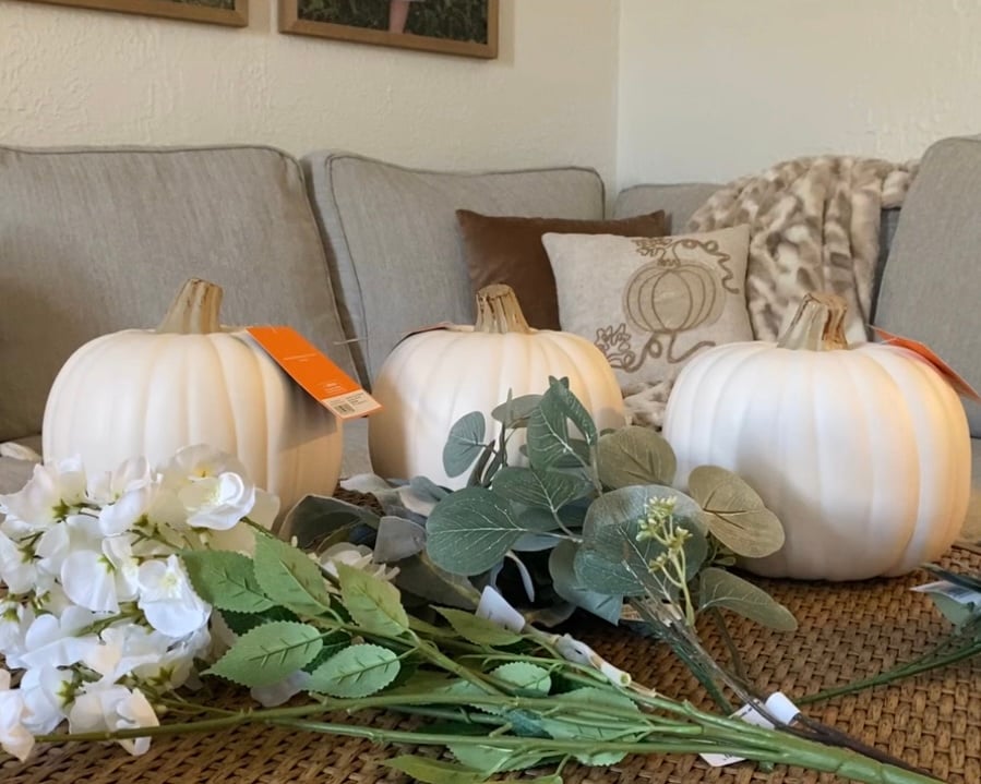 Three white faux pumpkins from Target with price tags still attached, displayed on a woven coffee table alongside faux white flowers and eucalyptus greenery before assembly for a DIY fall craft project.