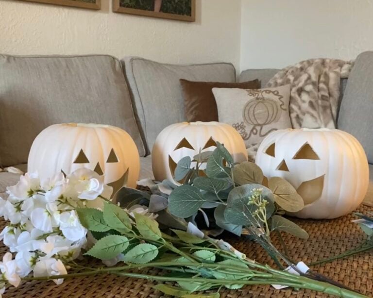 Three white faux pumpkins with carved jack-o’-lantern faces arranged on a woven coffee table with faux white flowers and greenery, styled in a cozy neutral living room for a fall DIY craft project.