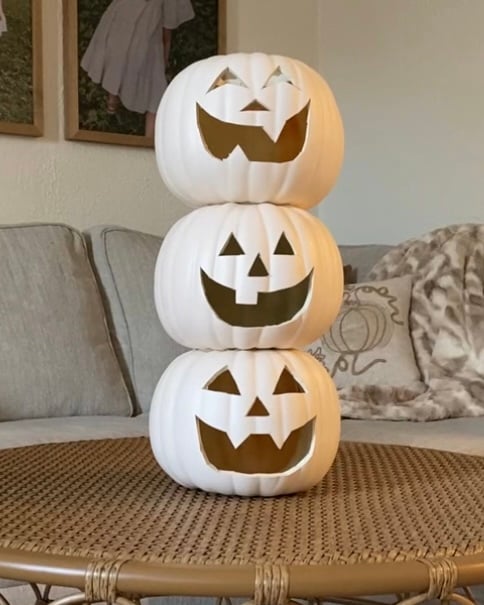 Three white faux pumpkins with carved jack-o’-lantern faces stacked vertically on a woven coffee table, showing the completed base of a DIY stacked pumpkin topiary before decoration.