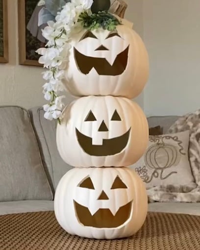 Stacked white jack-o’-lantern pumpkins decorated with faux white flowers and greenery on the top pumpkin, creating a finished DIY pumpkin topiary styled on a neutral woven coffee table.