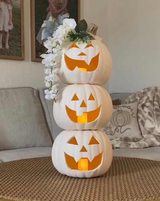 Finished DIY stacked pumpkin topiary with glowing tea lights inside each white jack-o’-lantern and floral accents on the top pumpkin, styled on a woven coffee table in a cozy neutral living room.