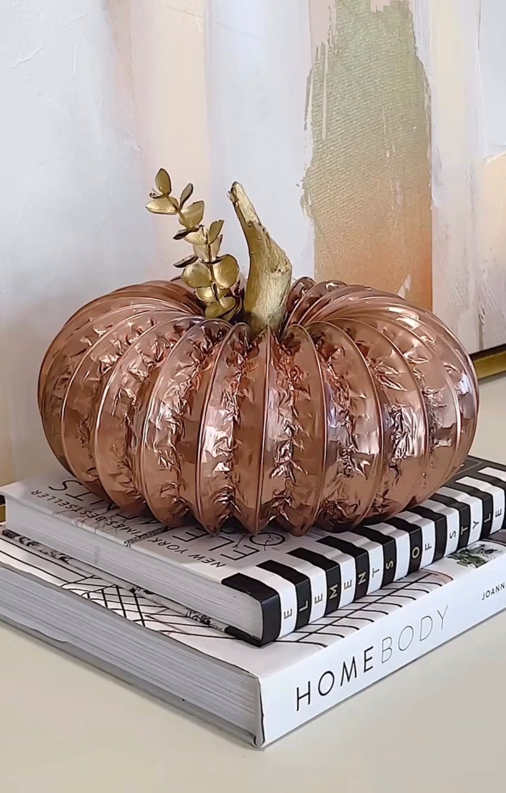 Rose gold glass pumpkin displayed on a stack of decorative coffee table books with a eucalyptus sprig, creating an elegant easy DIY fall decor idea for a living room or entryway.
