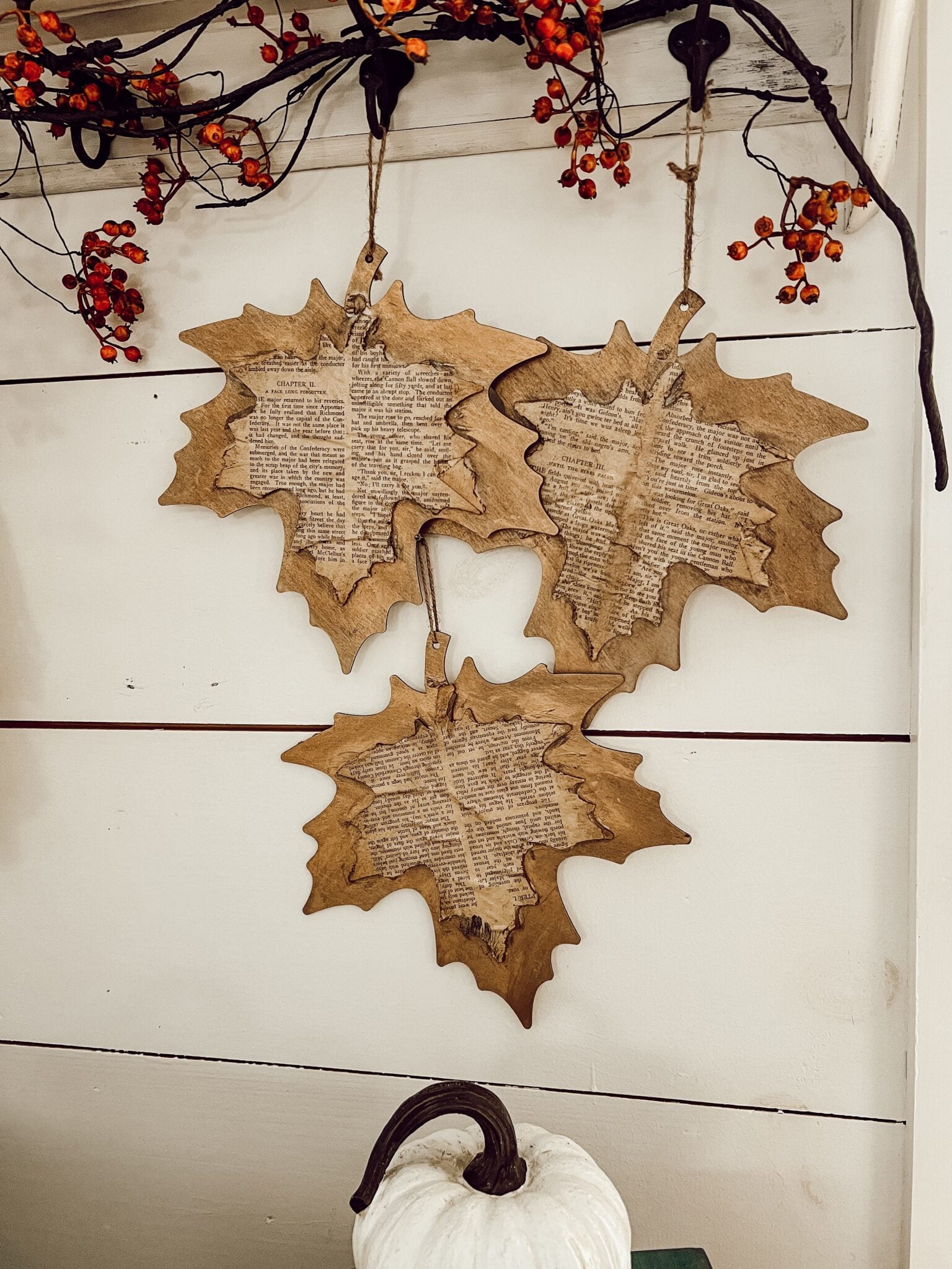 Wooden maple leaf ornaments with vintage book page decoupage hanging from twine beneath a berry garland for rustic easy DIY fall decor.