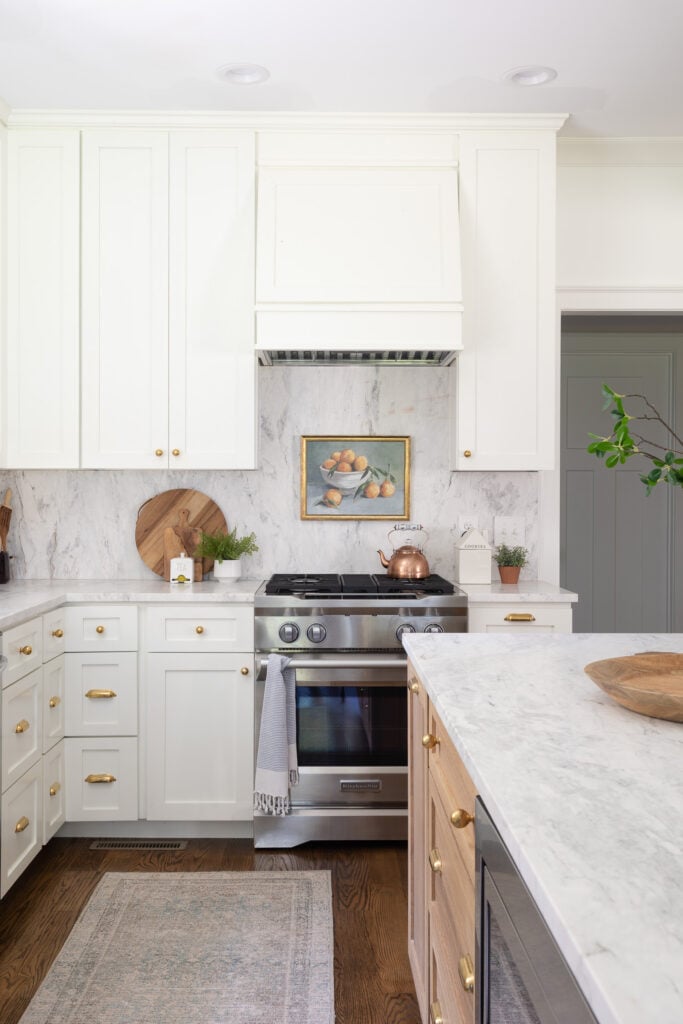 Popular kitchen backsplash idea featuring a full slab marble backsplash with soft gray veining, white shaker cabinets, brass hardware, and a stainless steel range.