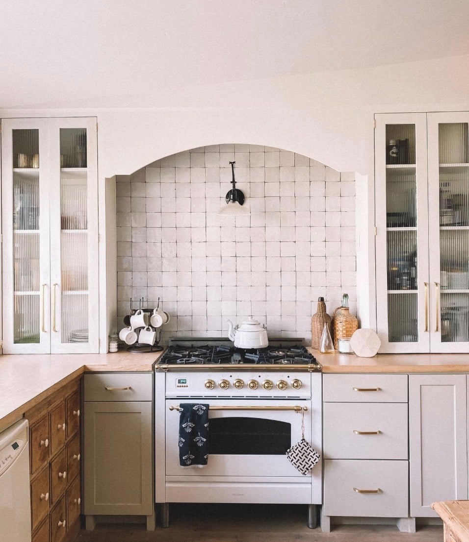 Popular kitchen backsplash idea featuring a cream zellige tile backsplash with an arched design, white cabinets, brass hardware, and a stainless steel range.