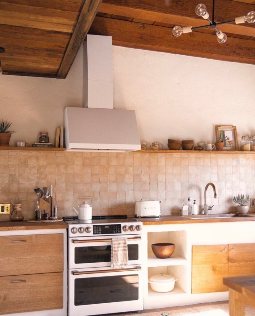 Popular kitchen backsplash idea featuring warm terracotta square tiles with natural color variation, paired with wood cabinets, open shelving, and rustic wood ceiling beams.