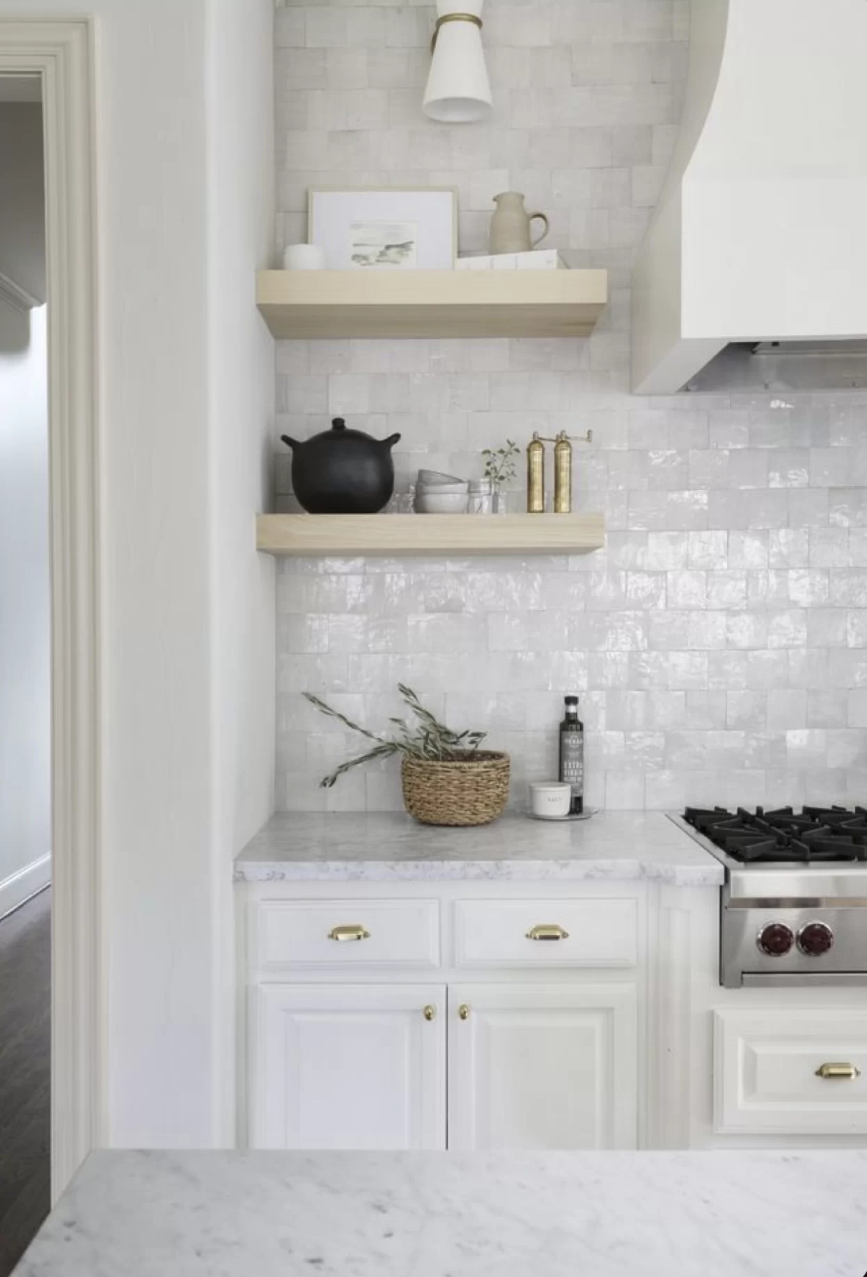 Popular kitchen backsplash idea featuring glossy white square tiles with subtle handmade texture, paired with white cabinets, marble countertops, brass hardware, and light wood floating shelves.