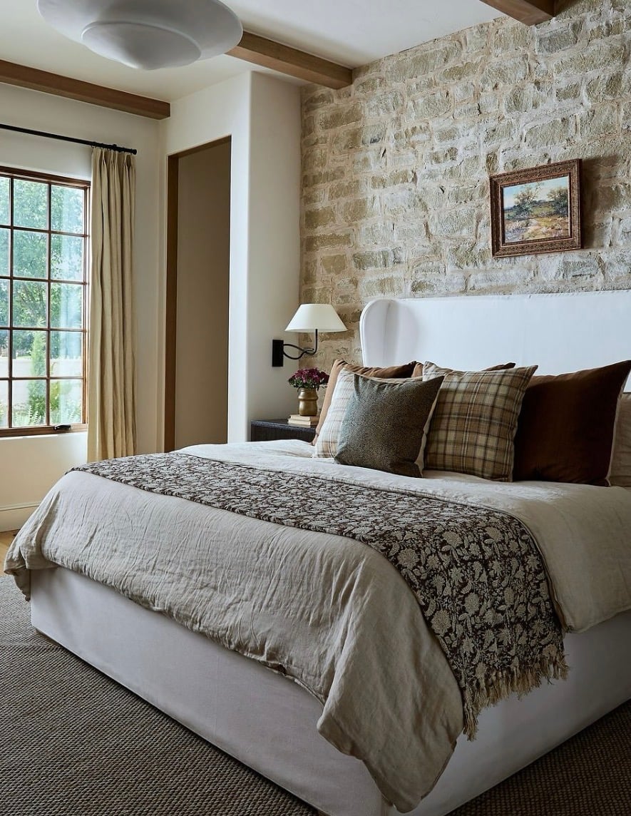 Natural stone accent wall behind a bed in a warm neutral bedroom with exposed wood ceiling beams, layered linens and rustic decor.