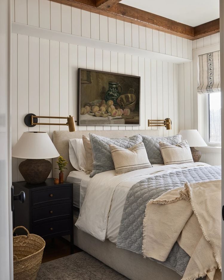 White vertical shiplap accent wall with rustic wood ceiling beams in a cozy farmhouse bedroom with layered neutral bedding.