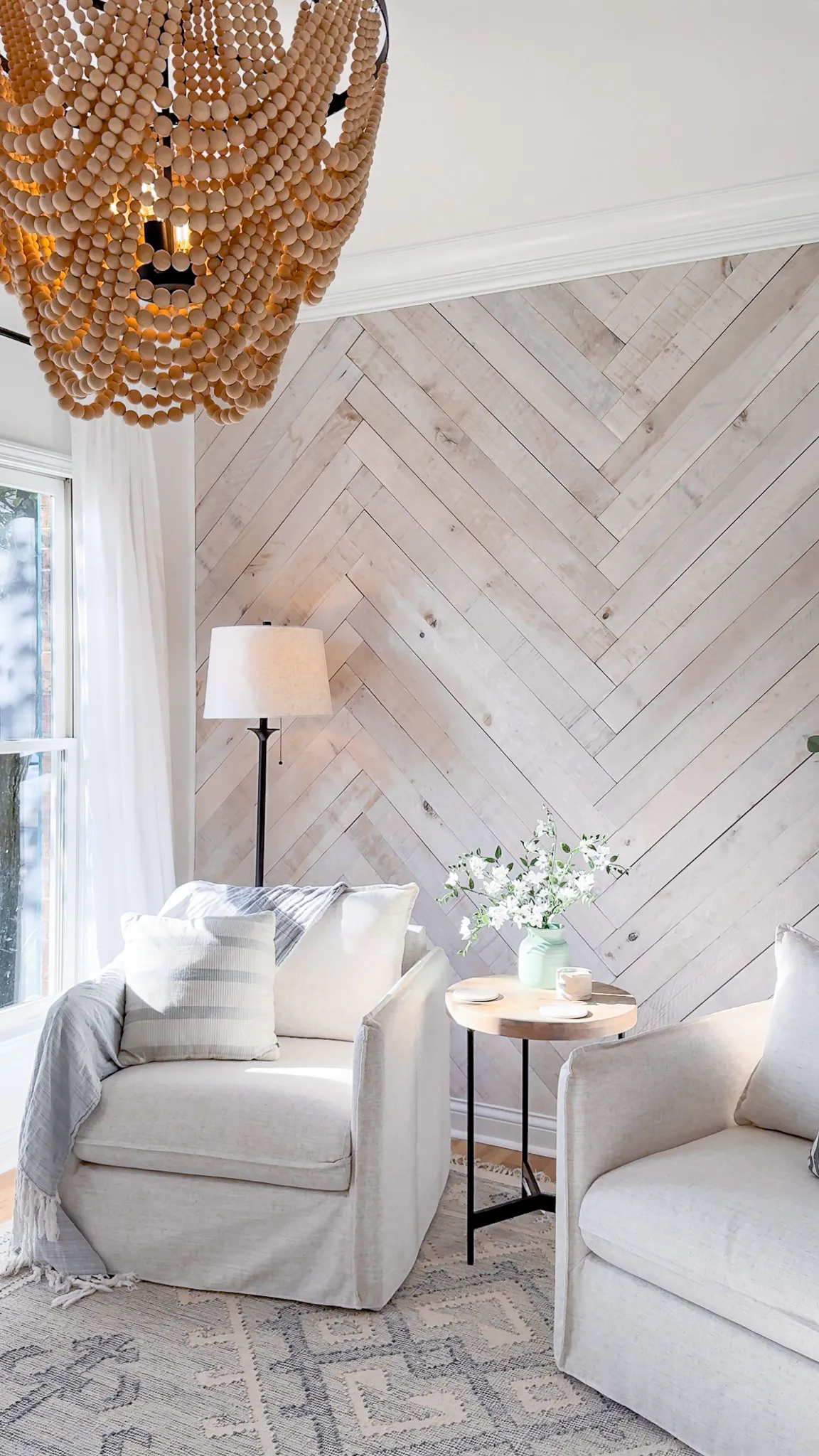 Whitewashed wood herringbone accent wall in a bright neutral living room with white chairs, a beaded chandelier and natural decor.