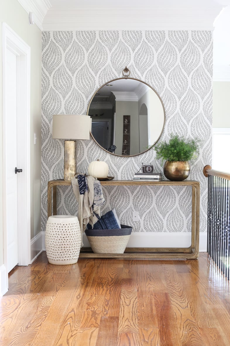 Gray and white patterned wallpaper accent wall in a bright hallway with a round mirror, console table, greenery and neutral decor.