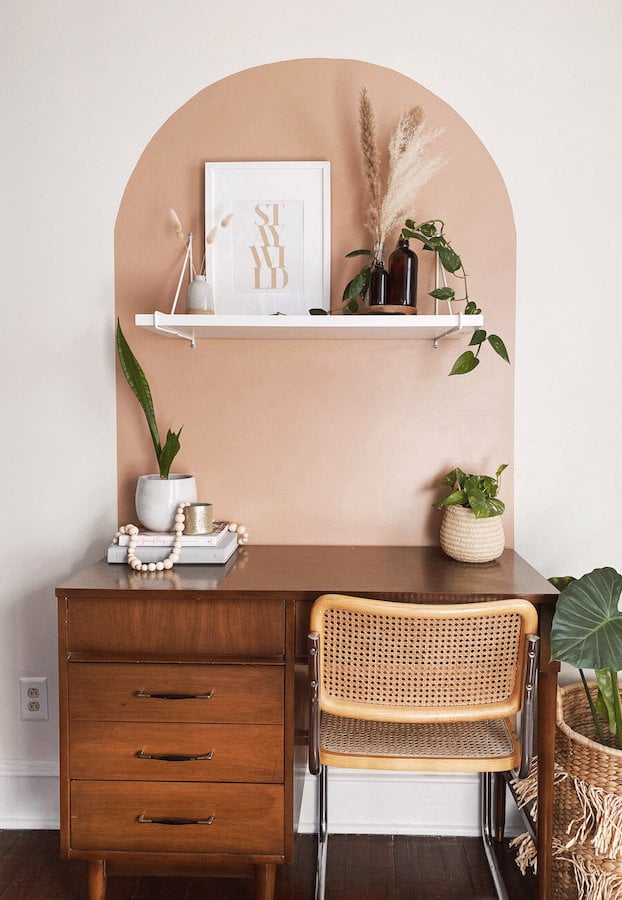 Soft blush pink painted arch accent wall framing a small home office with a vintage wood desk, cane chair, shelf and plants.