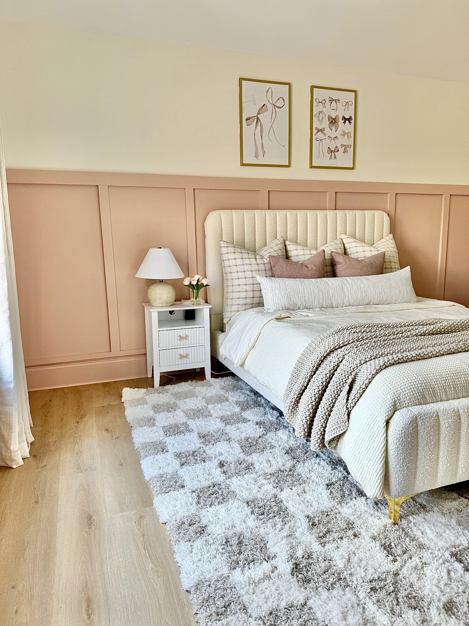 Soft dusty pink half-wall board and batten accent wall behind a bed in a cozy neutral bedroom with cream bedding and gold accents.