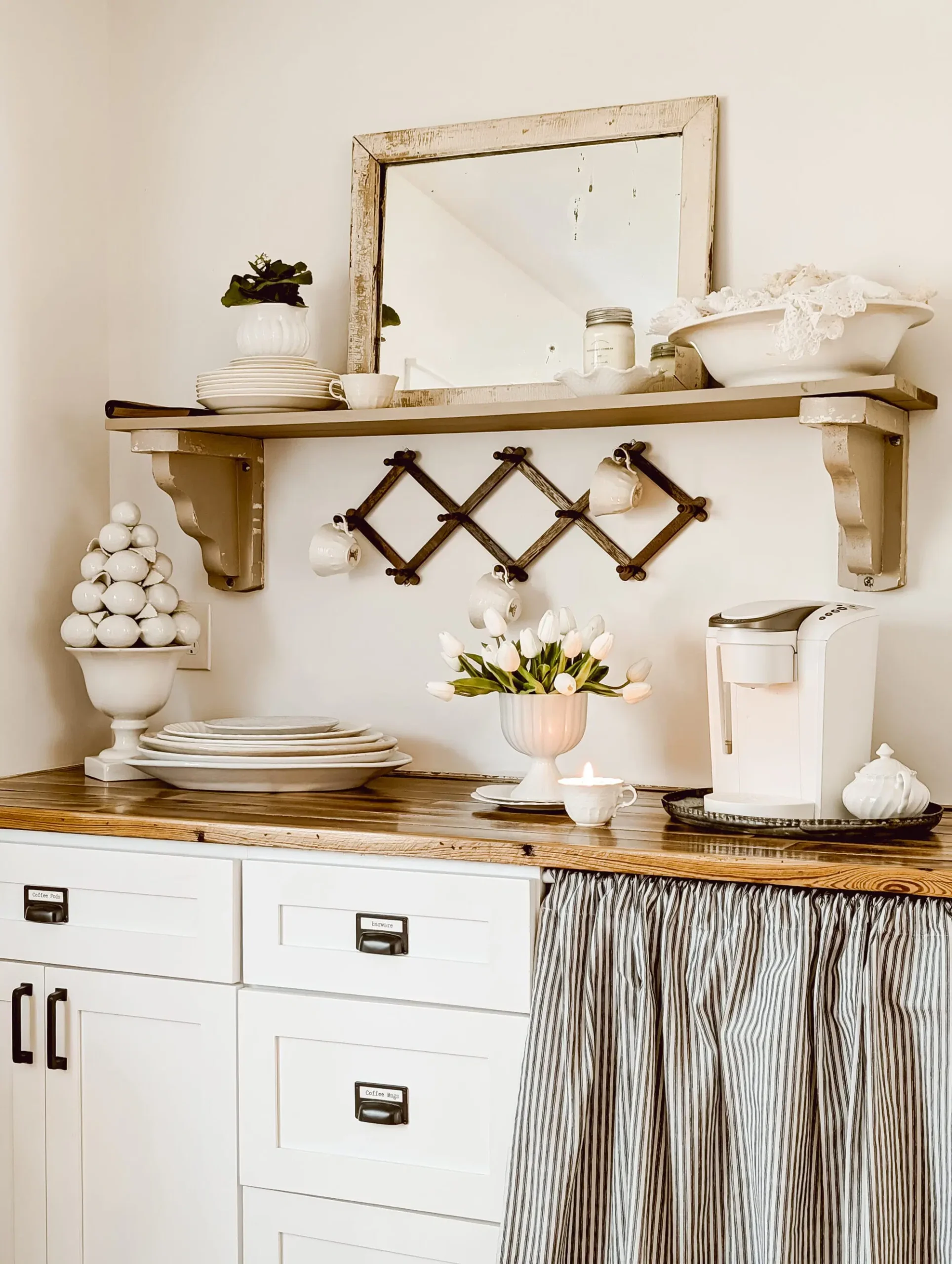 Rustic farmhouse kitchen coffee bar with a white Keurig, distressed wood countertop, vintage mirror, open shelving, hanging mugs, white tulips, and striped cabinet curtains.