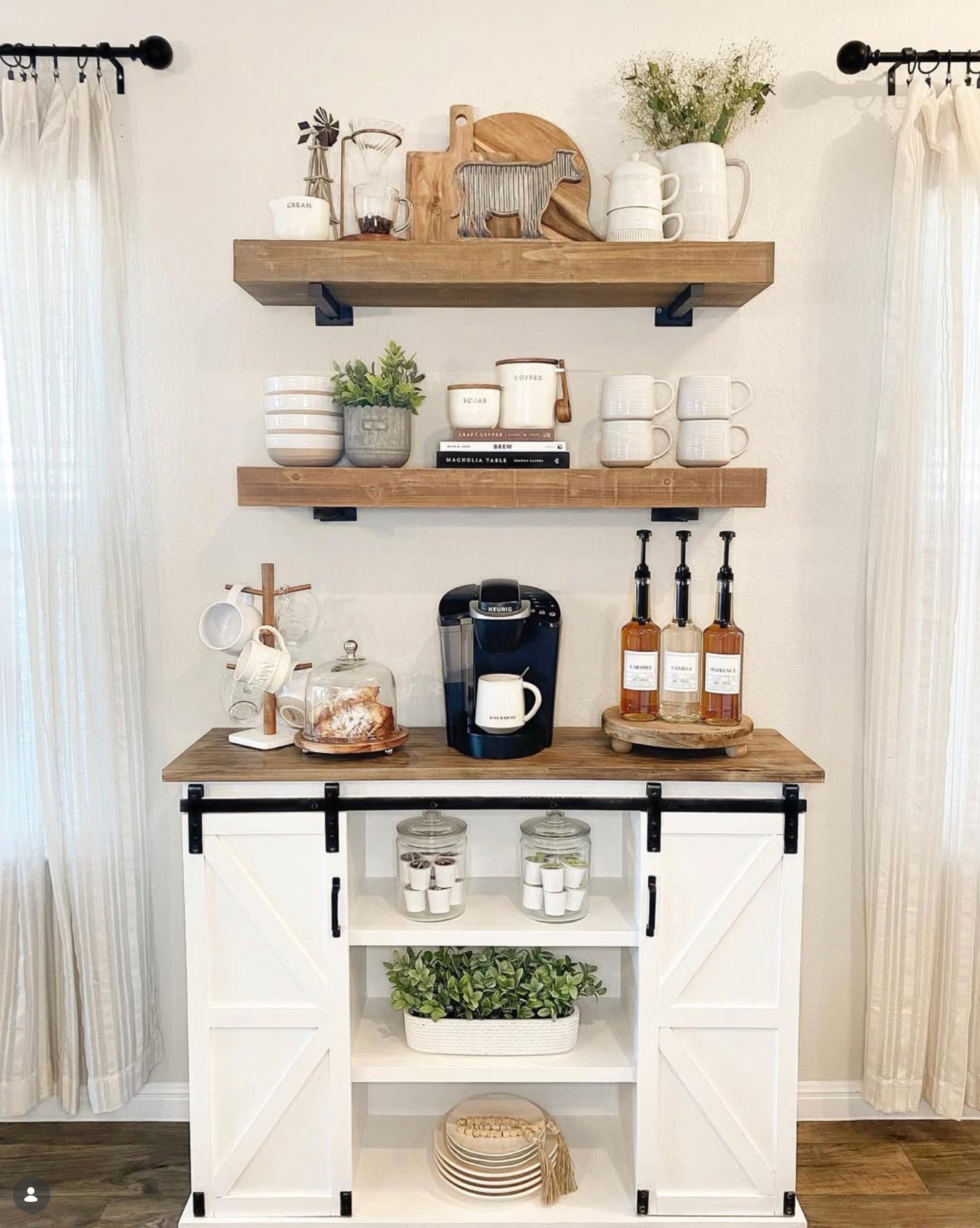 Farmhouse kitchen coffee bar with a white barn door cabinet, rustic wood floating shelves, black Keurig, coffee syrup bottles, hanging mugs, and greenery.