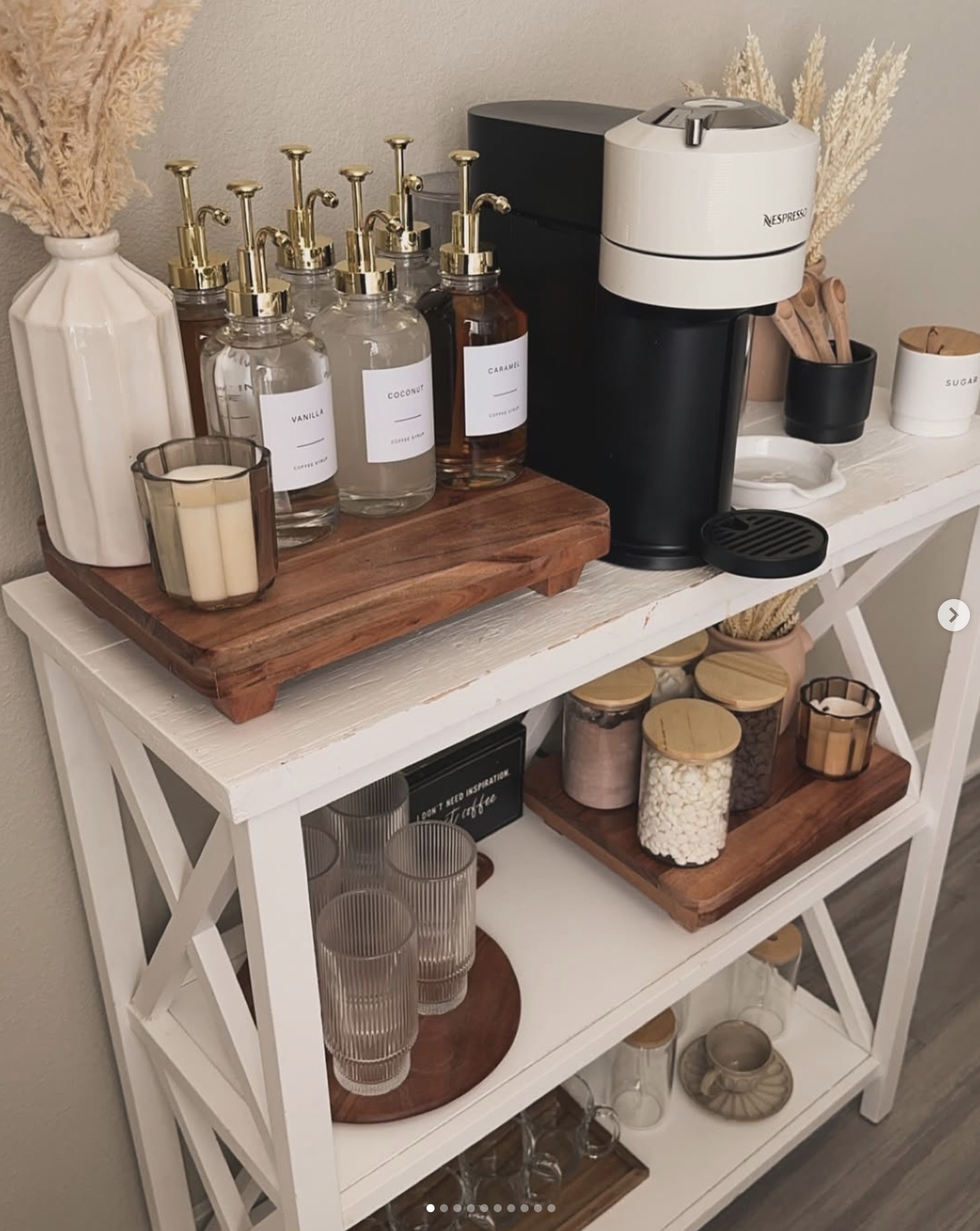 Boho kitchen coffee bar with a white console table, Nespresso machine, glass coffee syrup dispensers with gold pumps, wooden trays, pampas grass, and glass storage jars.