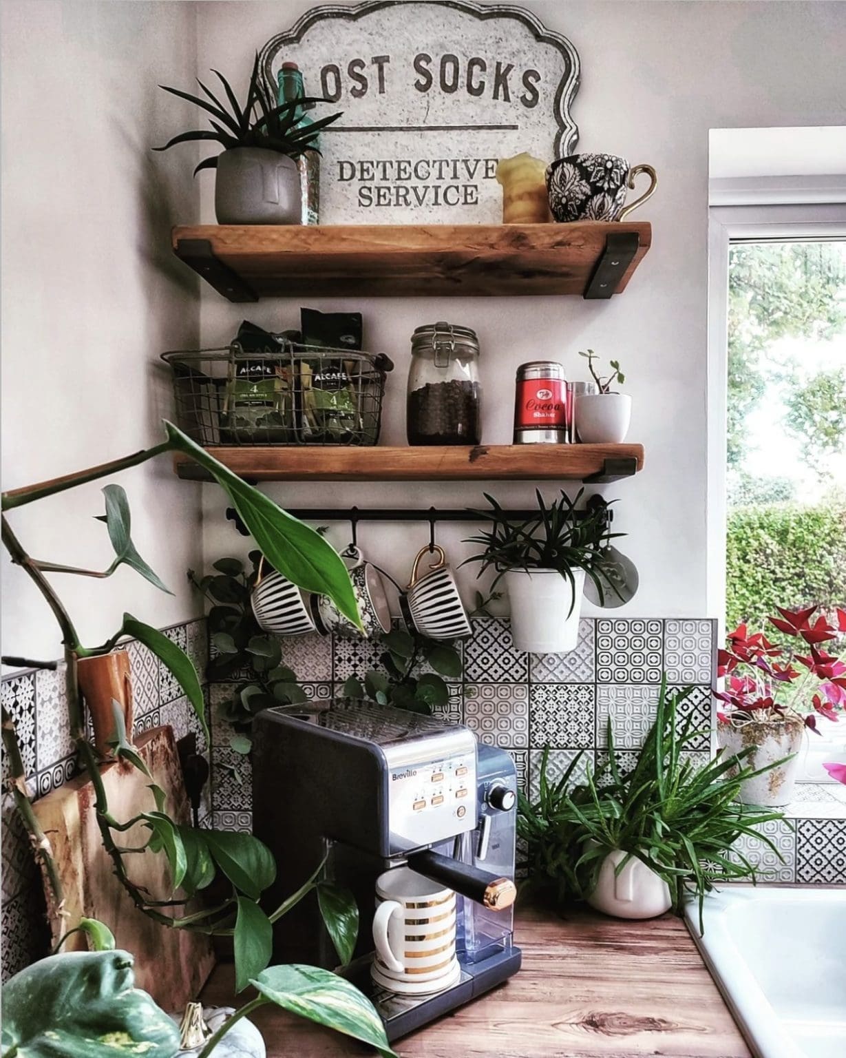 Nature-inspired kitchen coffee station with rustic wood floating shelves, patterned tile backsplash, hanging mugs, espresso machine, and lush green houseplants.
