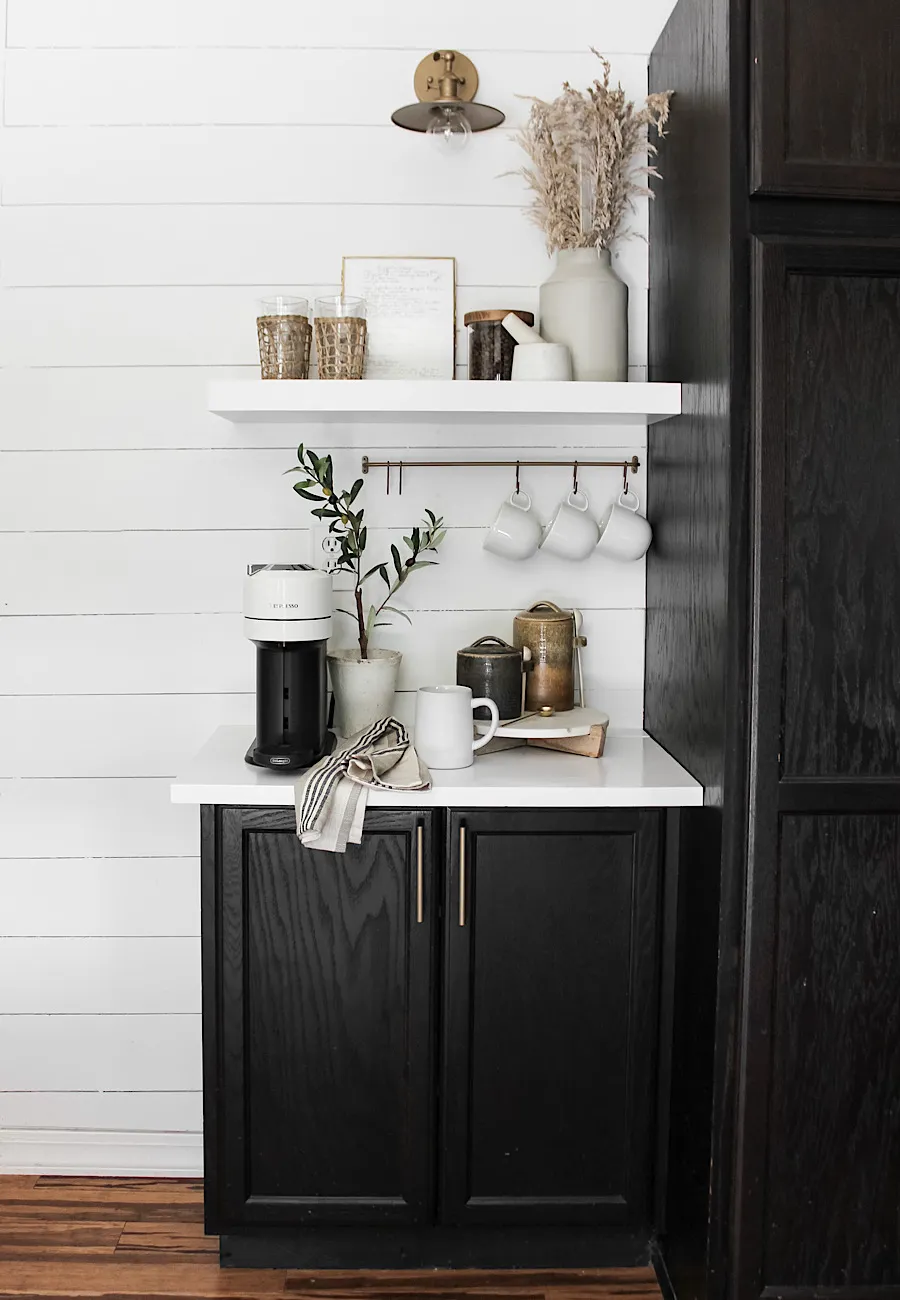 Simple and sleek kitchen coffee station with black cabinets, white shiplap walls, a floating shelf, brass mug rail, white Nespresso machine, and neutral decor.