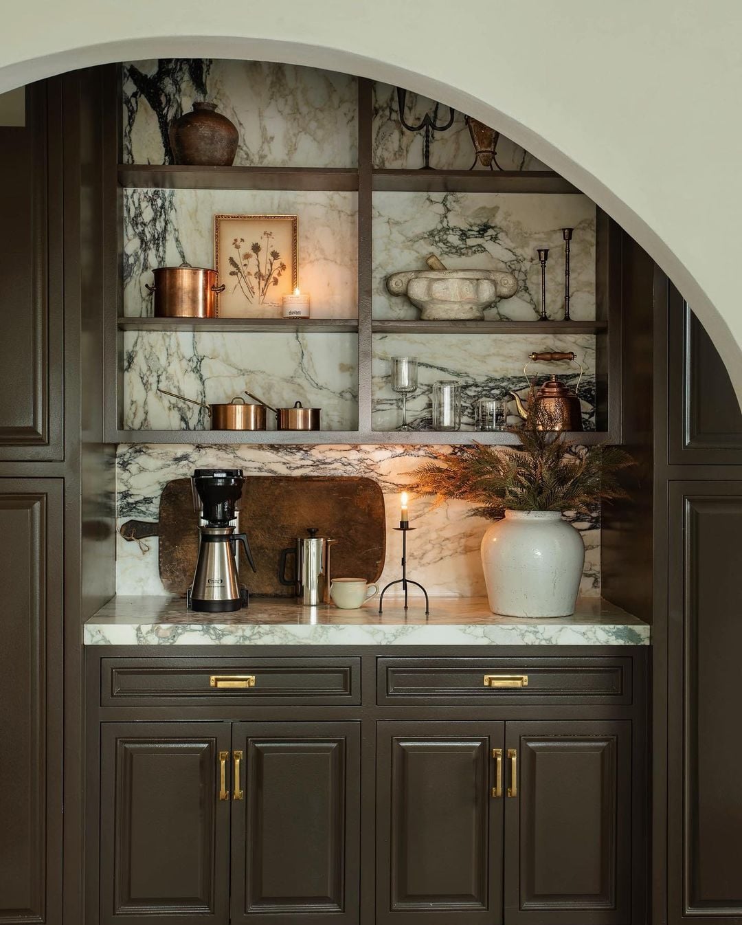 Luxurious and moody kitchen coffee bar with dark charcoal cabinetry, dramatic marble backsplash, arched alcove, brass hardware, open shelving, and copper accents.