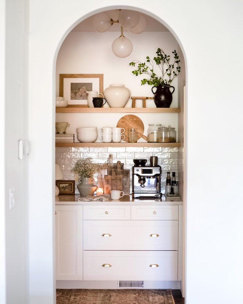 Kitchen pantry coffee bar tucked into an arched alcove with white cabinetry, natural wood floating shelves, glossy white tile backsplash, espresso machine, and brass hardware.