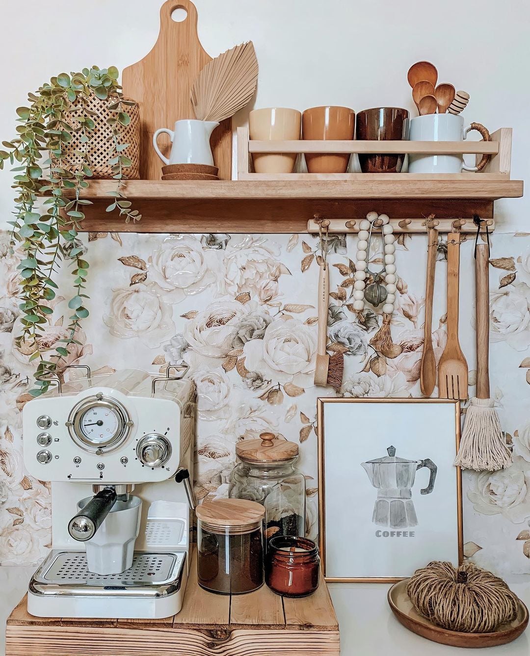 Boho kitchen coffee bar with a white espresso machine, floral wallpaper backsplash, rustic wood floating shelf, hanging wooden utensils, neutral ceramic mugs, and trailing greenery.