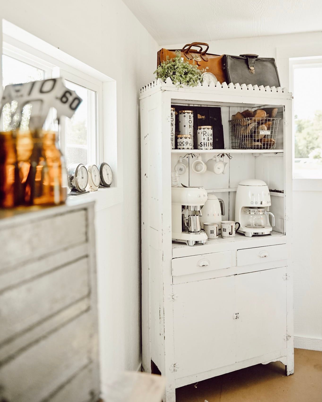 Repurposed vintage white hutch transformed into a farmhouse kitchen coffee bar with two white coffee makers, hanging mugs, open shelving, and antique decor.