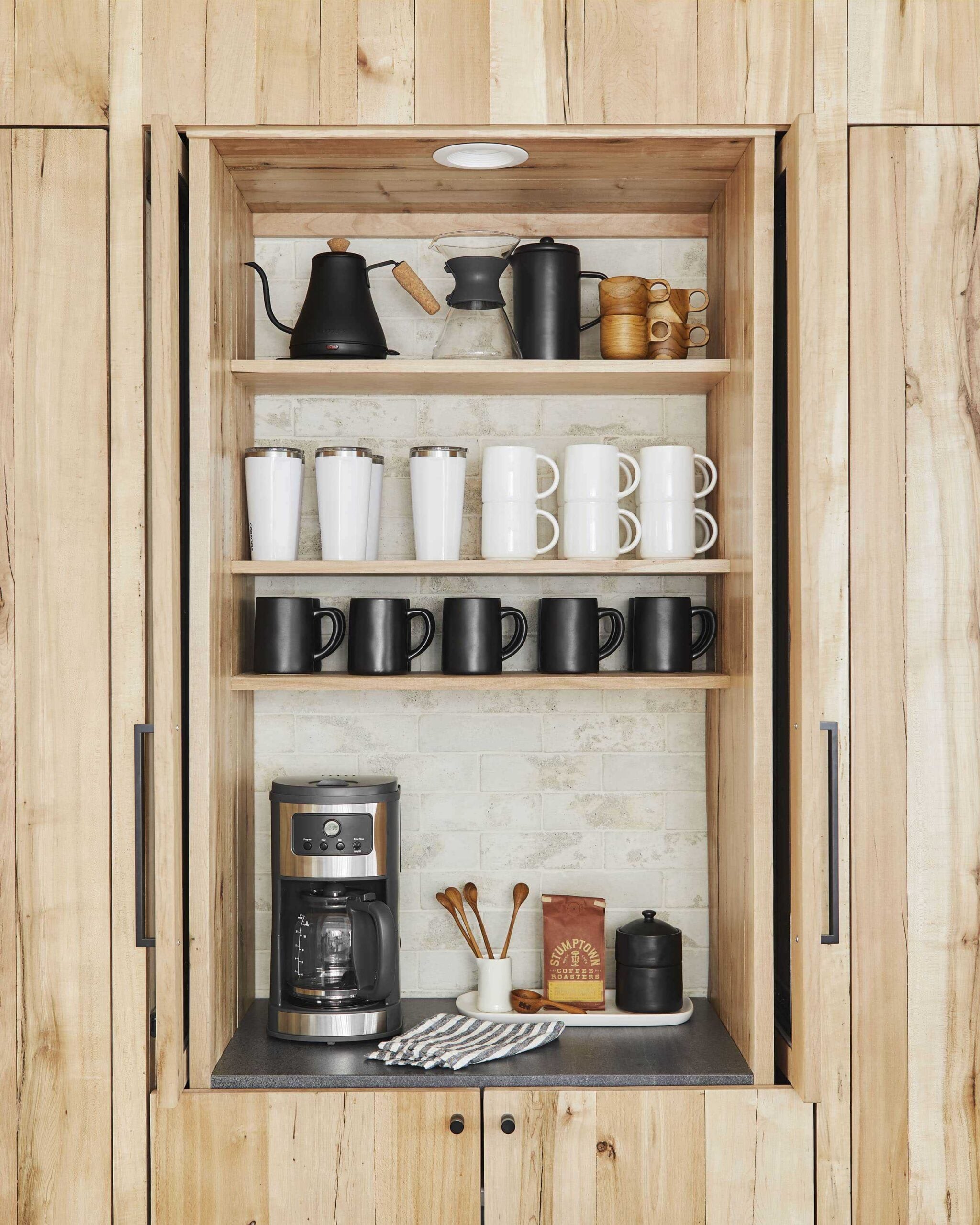 Built-in kitchen coffee bar with natural wood cabinetry, open shelving, white brick backsplash, black countertop, coffee maker, and neatly arranged black and white mugs.