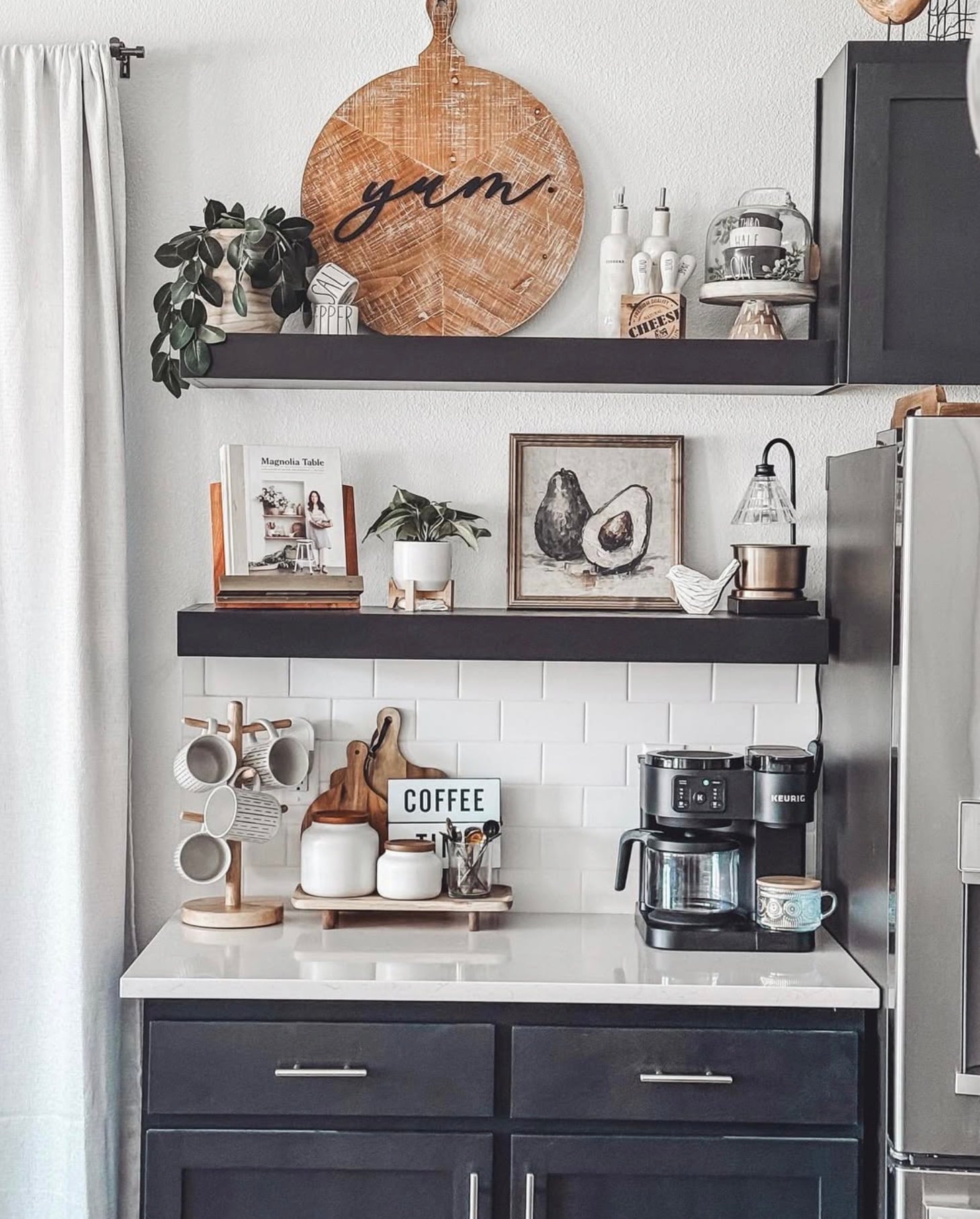 Modern farmhouse kitchen coffee station with black cabinets, white subway tile backsplash, black floating shelves, wooden mug rack, greenery, and a dual coffee maker.
