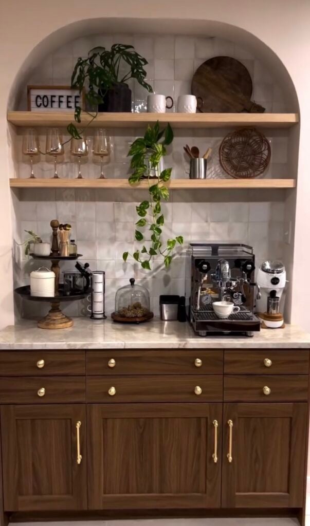 Built-in kitchen coffee bar with an arched alcove, warm wood cabinetry, marble countertop, white tile backsplash, floating shelves, espresso machine, and trailing greenery.