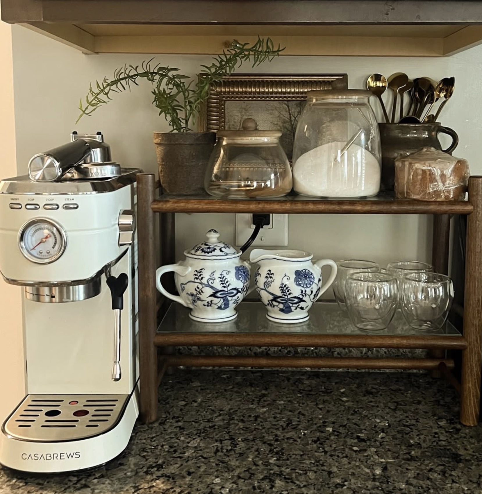 Vintage-inspired kitchen coffee station with a cream espresso machine, rustic wooden countertop shelves, blue and white floral ceramics, glass coffee cups, and antique-style decor.