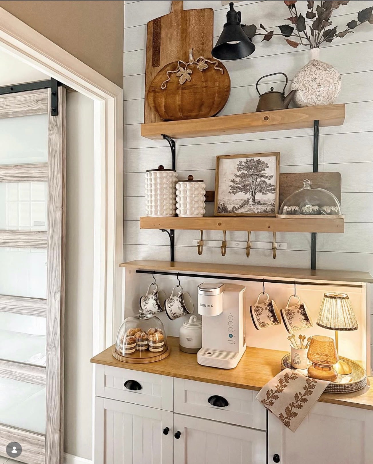 Fall country cottage kitchen coffee bar with white shiplap walls, rustic wood shelves, hanging floral mugs, white Keurig, vintage artwork, and warm autumn decor.