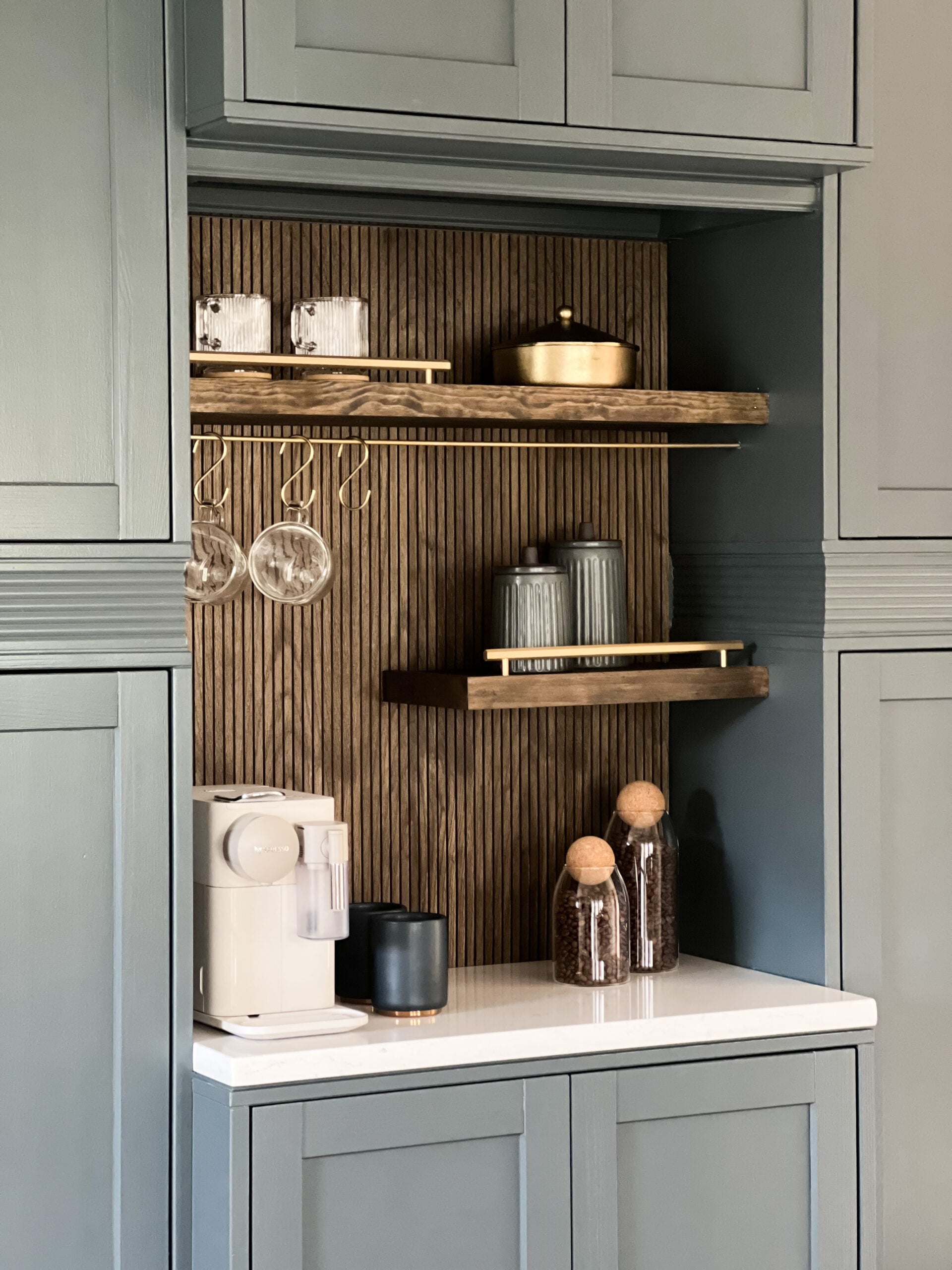 IKEA HAVSTA cabinet hack transformed into a built-in kitchen coffee station with sage green cabinetry, vertical wood slat backsplash, rustic floating shelves, brass mug hooks, and white Nespresso machine.