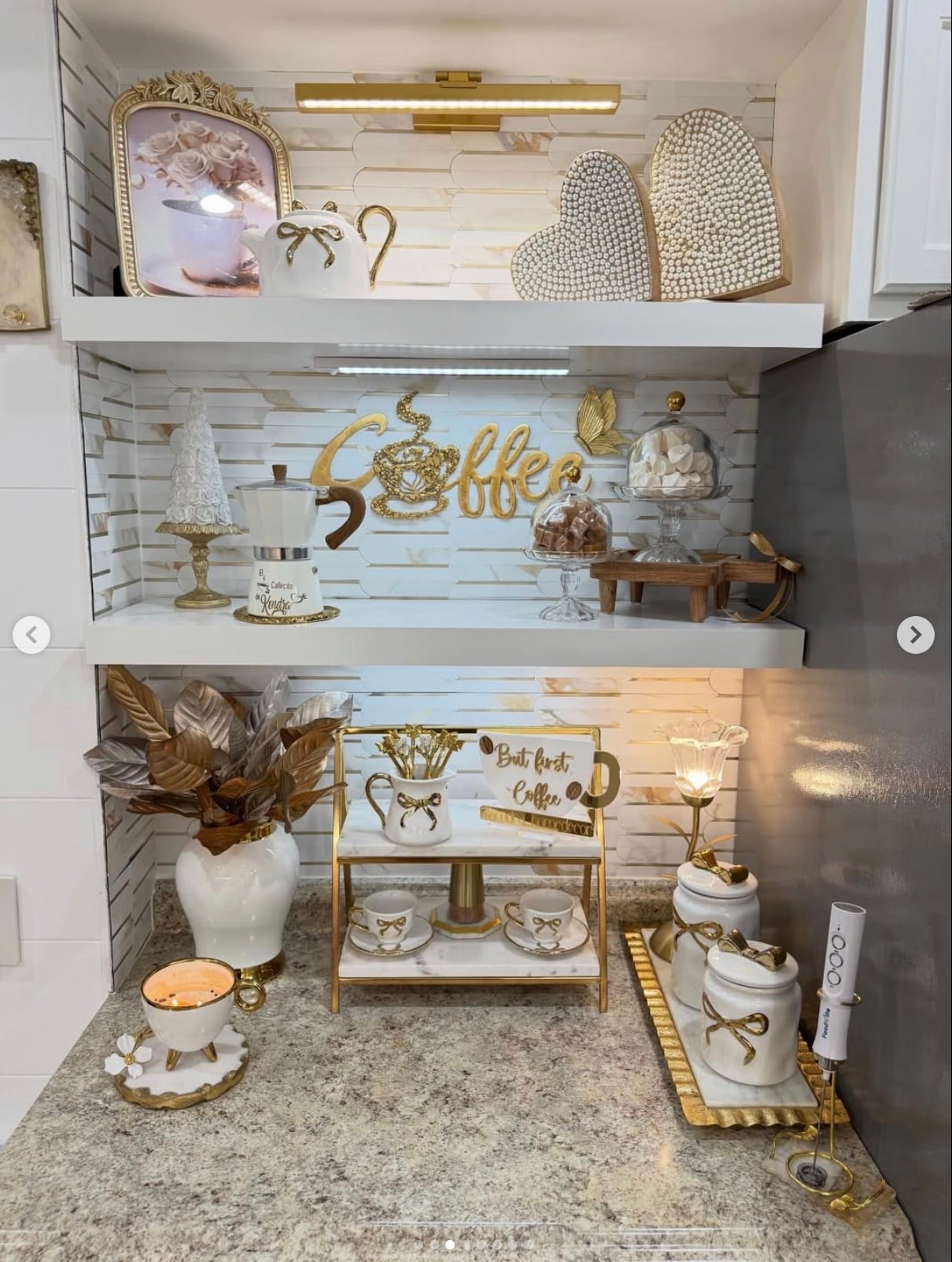 Glamorous kitchen coffee station with white floating shelves, gold coffee decor, marble and gold tiered tray, decorative bow canisters, framed floral artwork, and warm accent lighting.