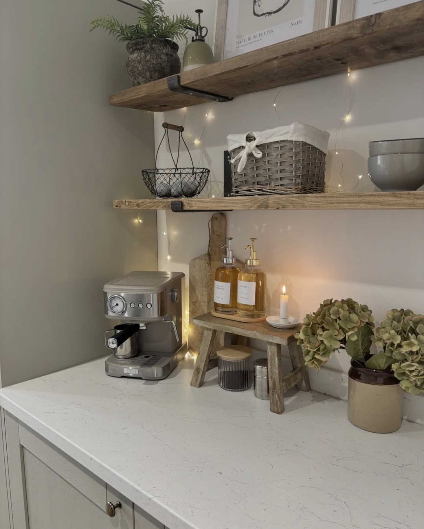 Rustic kitchen coffee station with reclaimed wood floating shelves, a weathered wood riser, glass coffee syrup dispensers, espresso machine, woven baskets, and warm fairy lights.