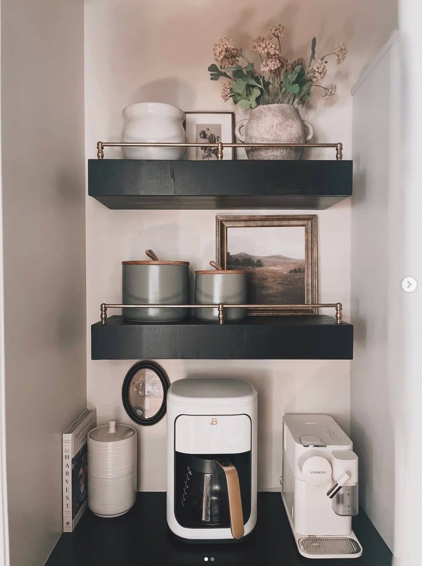 Elegant kitchen coffee station with dark charcoal floating shelves, brass gallery rails, matching coffee canisters, framed artwork, white coffee makers, and soft pink florals.