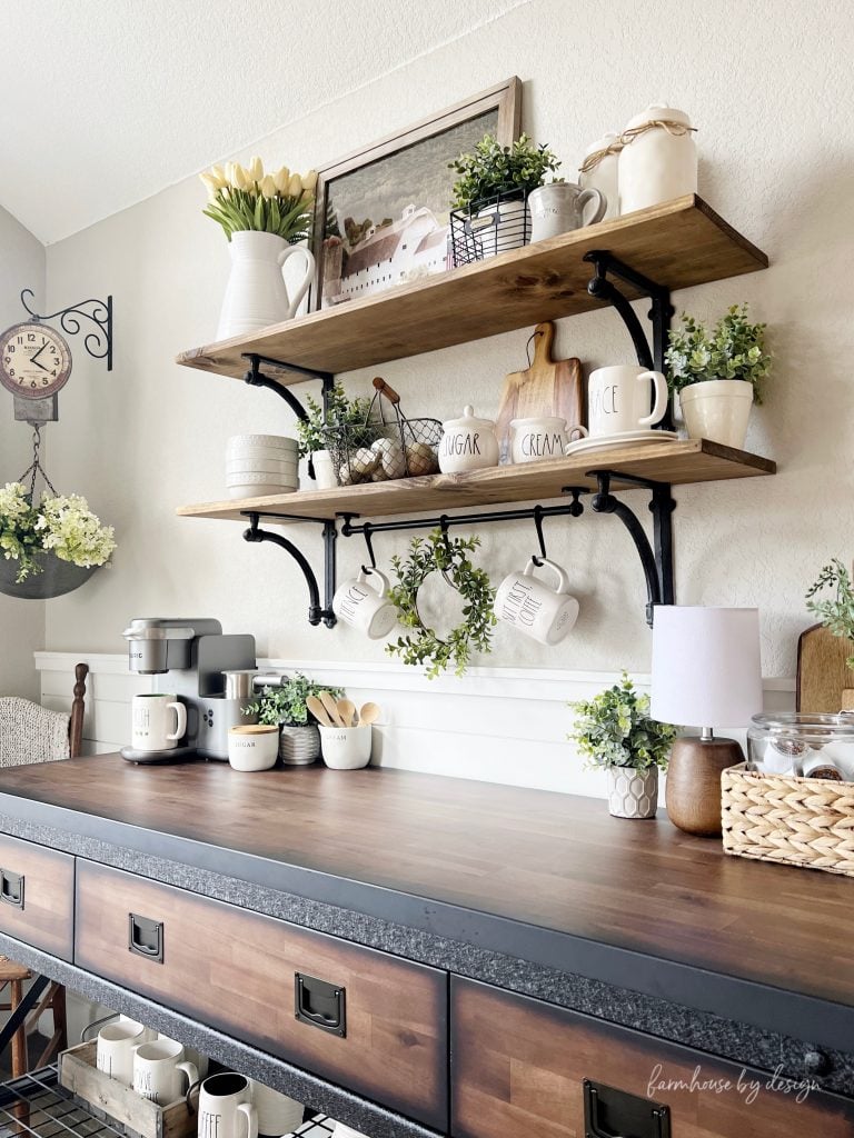 Rustic farmhouse kitchen coffee bar with DIY wood floating shelves, black metal brackets, hanging coffee mugs, greenery, white ceramic canisters, and a long wooden countertop.