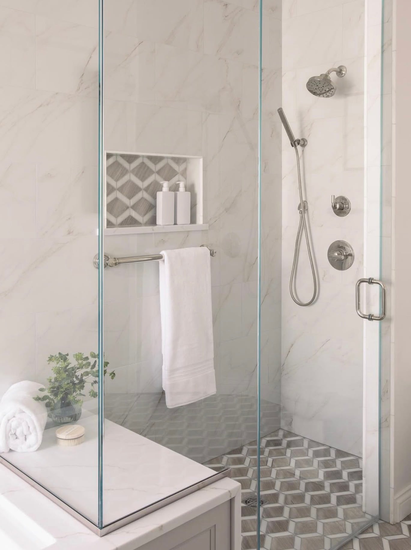 Modern walk-in tile shower with large white marble-look wall tiles, gray geometric mosaic floor tiles, matching tiled shower niche, frameless glass enclosure, and brushed nickel fixtures.