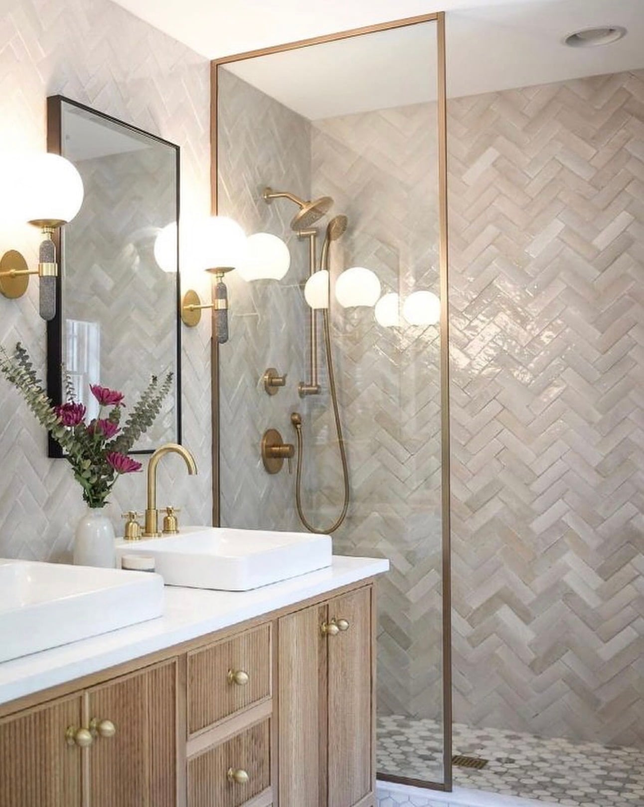 Modern walk-in shower with glossy beige herringbone wall tiles, small marble mosaic floor tiles, brushed brass shower fixtures, and a glass enclosure with gold trim.