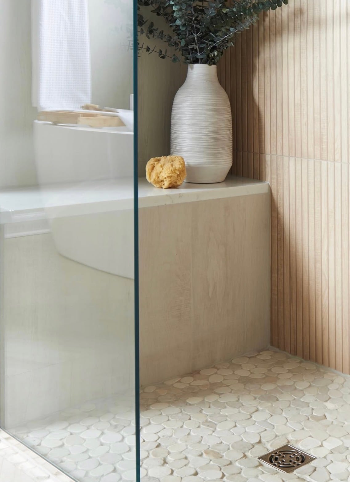 Nature-inspired walk-in shower with vertical light wood-look wall tiles, cream pebble stone shower floor, frameless glass enclosure, and built-in bench beside a white soaking bathtub.