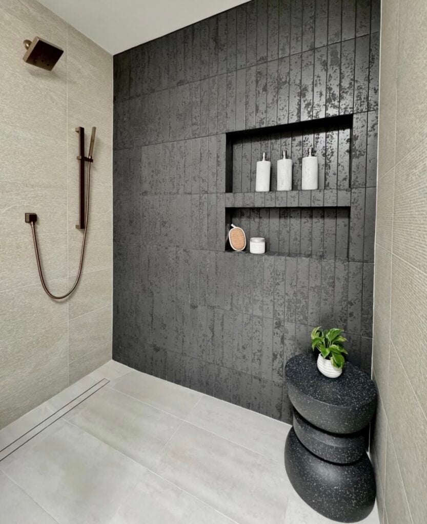 Modern walk-in shower with dark charcoal textured vertical tile accent wall, large light gray porcelain wall and floor tiles, two recessed shower niches, bronze shower fixtures, and black stone stool.