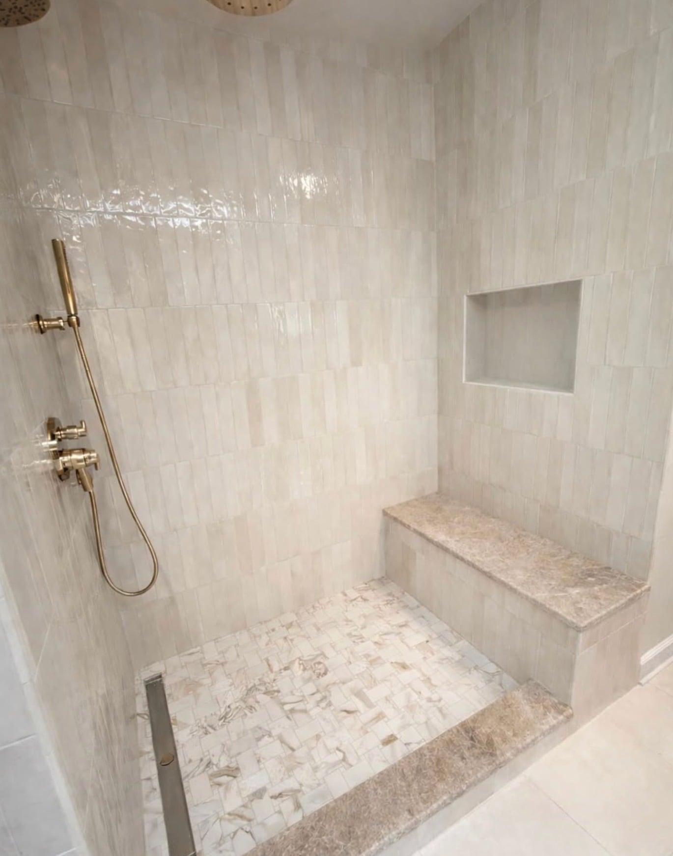 Luxury walk-in tile shower with brushed gold rainfall showerhead, matching gold handheld shower fixtures, glossy cream vertical wall tiles, marble mosaic flooring, and built-in shower bench.
