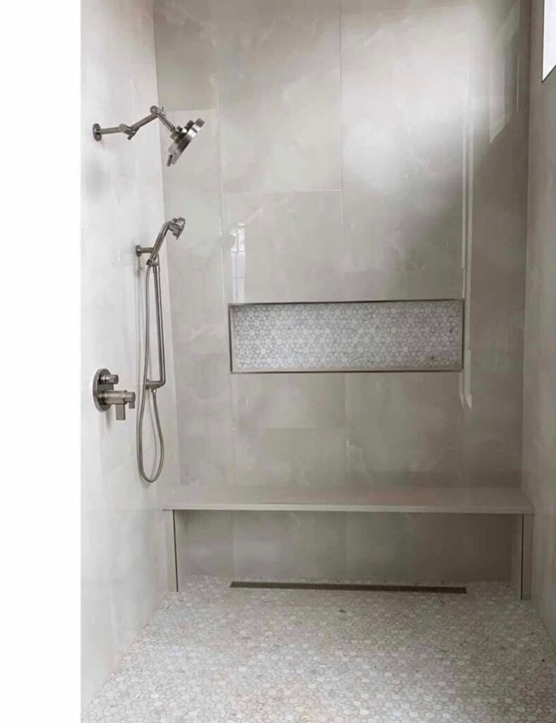 Modern walk-in tile shower with large light gray marble-look wall tiles, white and gray penny mosaic tile flooring, matching mosaic shower niche, built-in shower bench, and brushed nickel rainfall showerhead with handheld sprayer.