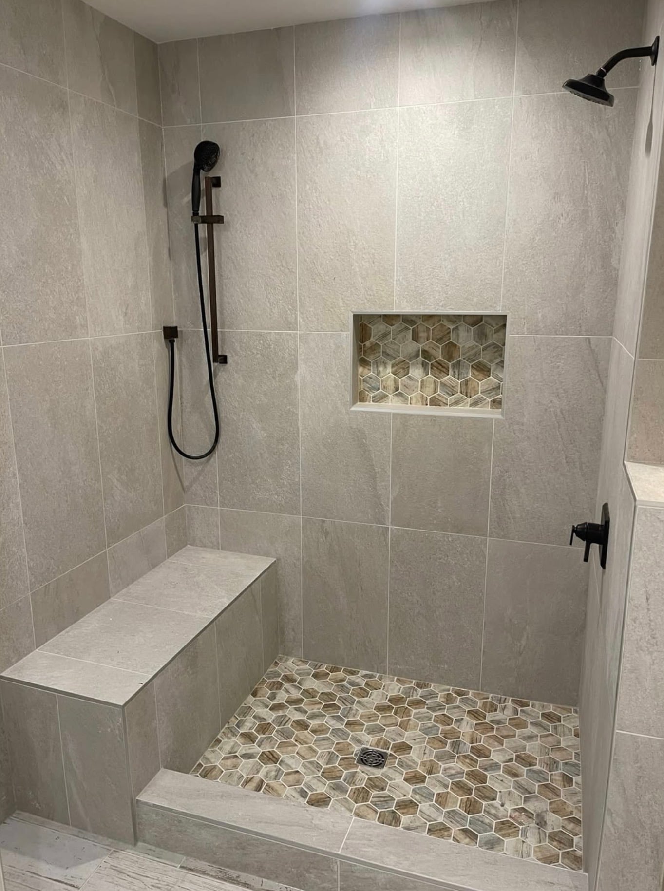 Modern walk-in tile shower with large light gray stone-look wall tiles, brown and beige hexagon mosaic shower floor, matching hexagon tile shower niche, built-in tiled bench, and matte black shower fixtures.