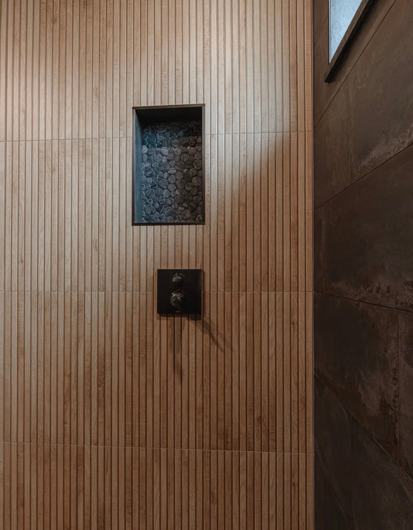 Modern shower with vertical fluted wood-look wall tiles in warm natural brown, dark pebble stone mosaic shower niche, black shower fixtures, and large dark charcoal stone-look accent wall tiles.