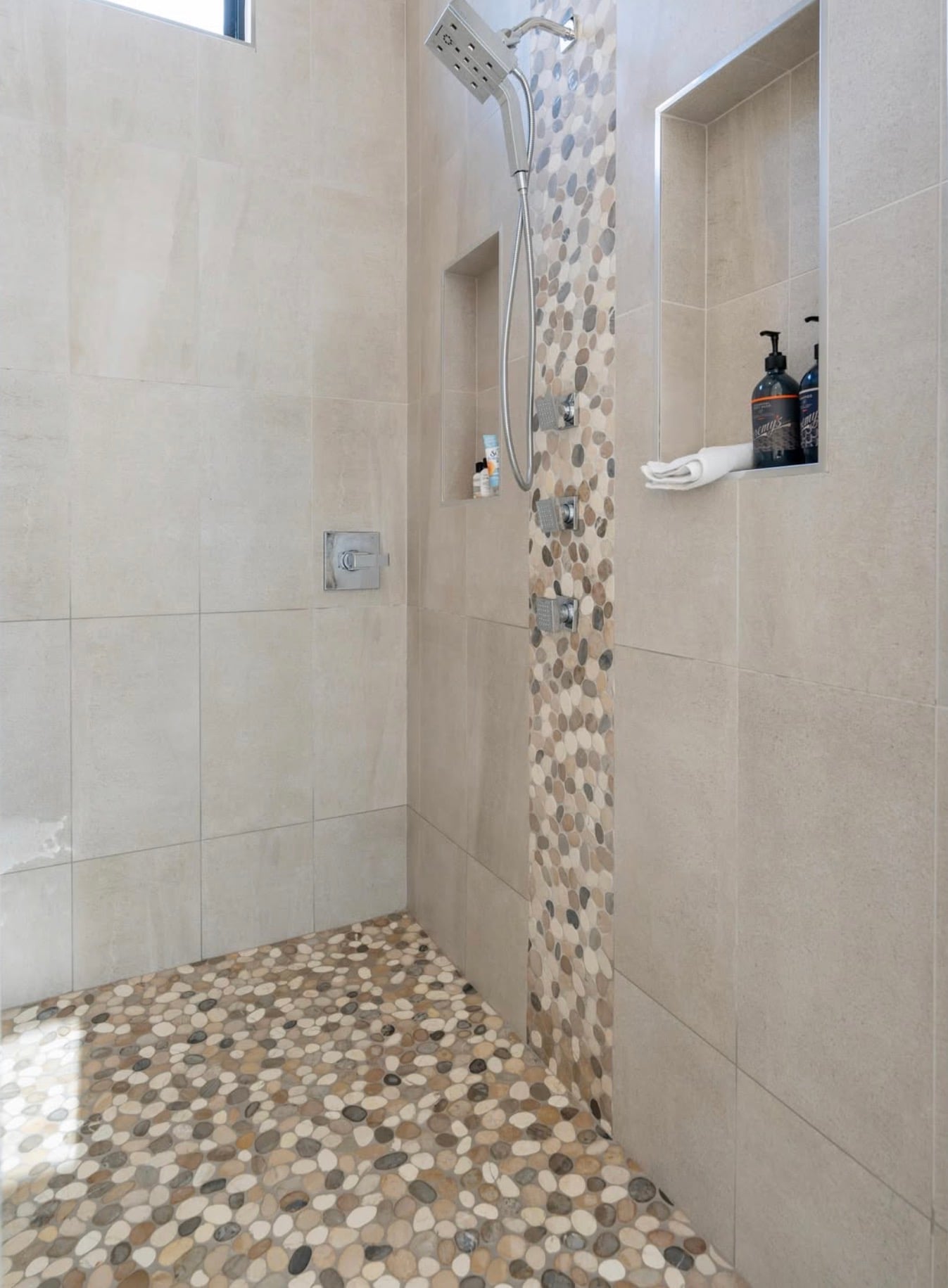 Modern walk-in shower with multicolored river rock pebble tile flooring, matching vertical pebble stone accent strip, large beige porcelain wall tiles, recessed shower niches, and chrome shower fixtures.