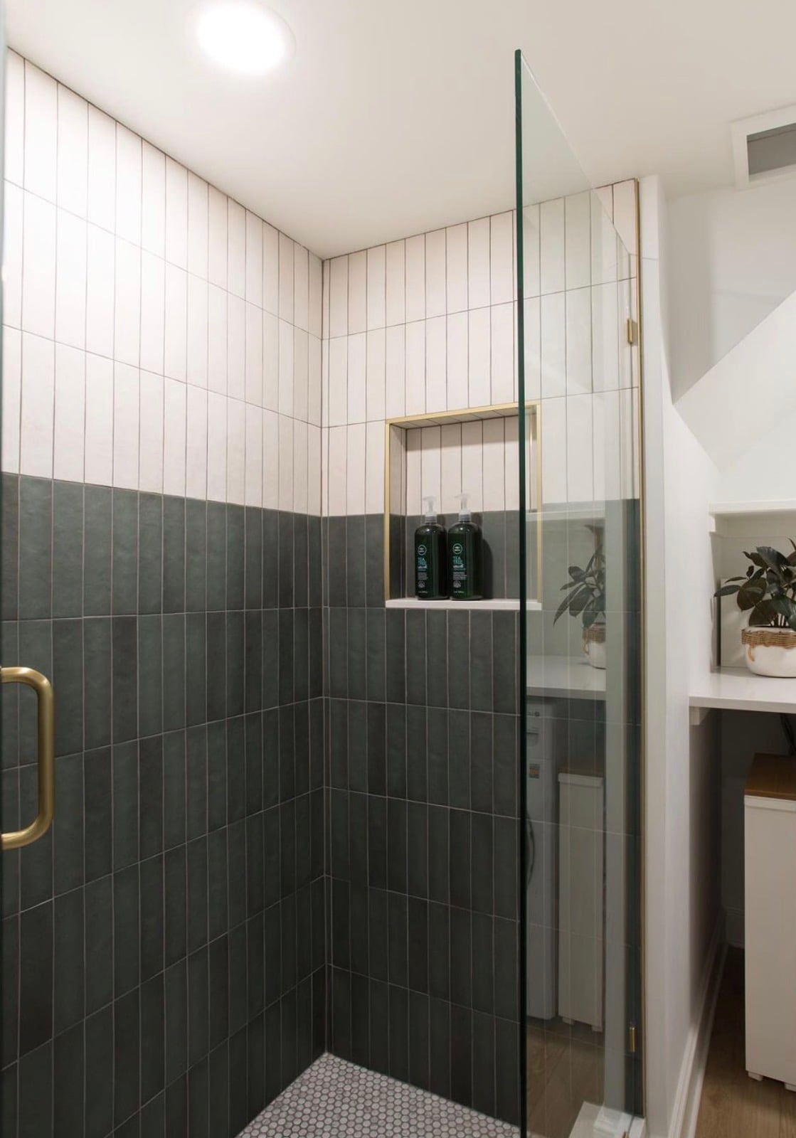 Modern dual-color tile shower with white vertical stacked subway tiles on the upper walls, dark charcoal green vertical tiles on the lower walls, gold-trimmed shower niche, brass glass door handle, and white penny tile flooring.